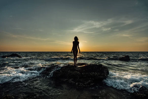 La silueta de la muchacha hermosa al atardecer junto al mar, sobre la piedra de mar en el bañador. Olas, espuma . — Foto de Stock