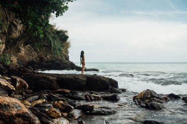 Okyanus kıyısında bir kaya üzerinde sarı bikini duran güzel genç kadın