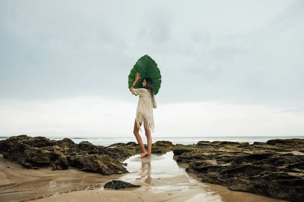 Retrato joven morena mujer con una hoja de palma de pie en la costa de fondo mar — Foto de Stock