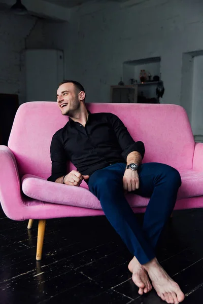 男モデルはロフトのインテリアのピンクのソファの上に座っています。黒の t シャツ、時計、青いズボンのビジネスマン. — ストック写真