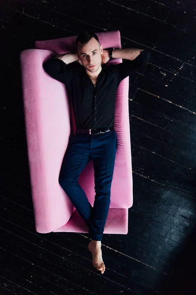 Model mężczyzna siedzi na różowy sofa strych wnętrza. Biznesmen w czarnej koszulce i zegarek, niebieskie Spodnie. — Zdjęcie stockowe