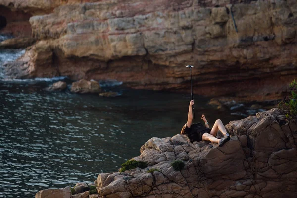 Mujer haciendo selfie usando selfie stick. joven se encuentra en una roca, sobre un fondo de rocas y lago . — Foto de Stock