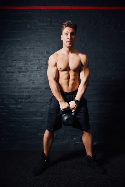 Jonge gespierde man trainen met kettlebells. man met naakte torso op donkere achtergrond. — Stockfoto