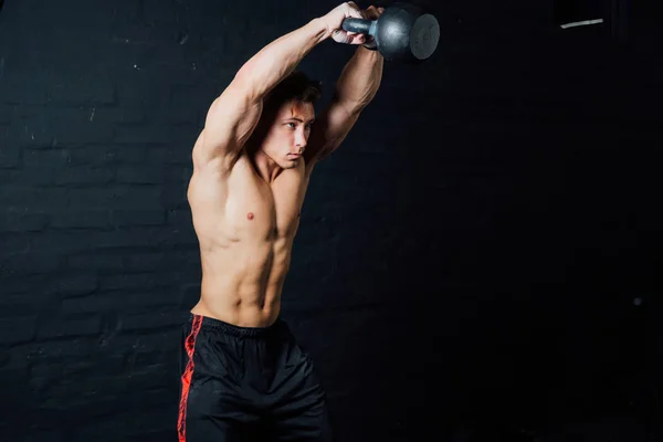 Молодой мускулистый человек тренируется с гирями. Человек с обнаженным туловищем на темном фоне . — стоковое фото