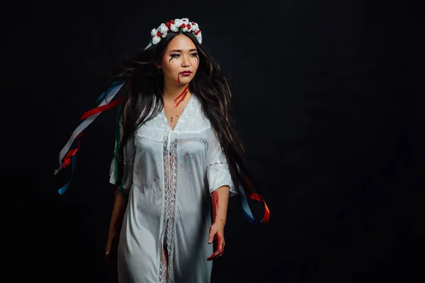 Portrait of a bloody beautiful Asian girl on Halloween. White dress-robe, ethnic wreath of flowers. — Stock Photo, Image