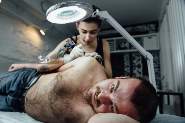 在沙发上躺着的人的正面肖像, 纹身沙龙的客户在过程中制作纹身的肩膀 . — 图库照片