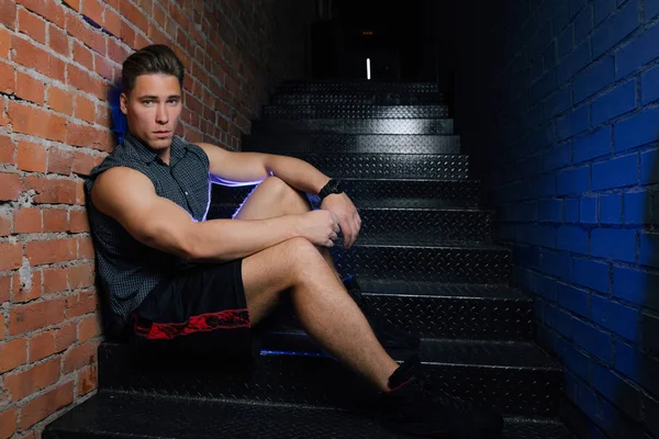Fitness model man in shirt sleeveless posing sitting on stairs in fitness club loft. Red brick wall.