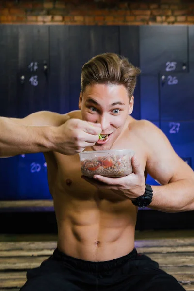 Modelo de fitness de un culturista comiendo comida de un contenedor de plástico en un vestuario masculino . —  Fotos de Stock