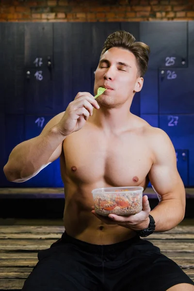 Fitness model kulturista člověk jíst jídlo z plastové nádoby v mužské šatně. — Stock fotografie