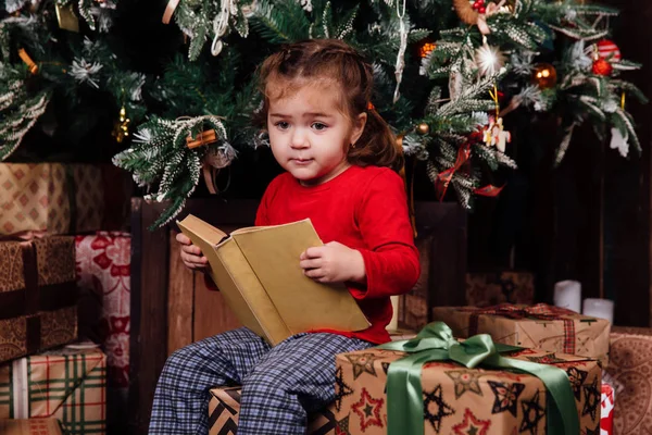 Küçük kız tarafından bir Noel ağacı hikayeleri bir kitap okuyor. Kırmızı gömlek pijama. Hediye kutuları üzerinde oturur. — Stok fotoğraf