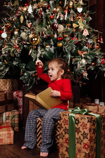 Küçük kız tarafından bir Noel ağacı hikayeleri bir kitap okuyor. Kırmızı gömlek pijama. Hediye kutuları üzerinde oturur. — Stok fotoğraf