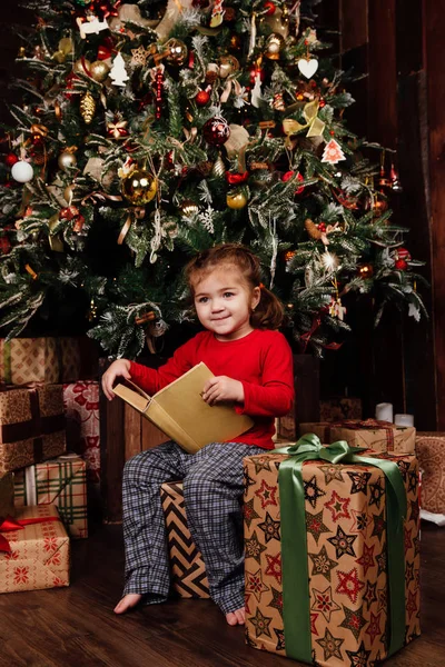 Küçük kız tarafından bir Noel ağacı hikayeleri bir kitap okuyor. Kırmızı gömlek pijama. Hediye kutuları üzerinde oturur. — Stok fotoğraf