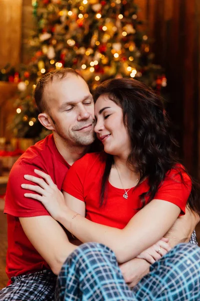 Casal adulto casado em camisetas vermelhas e pijamas abraça sentado no chão contra a árvore de Natal . — Fotografia de Stock