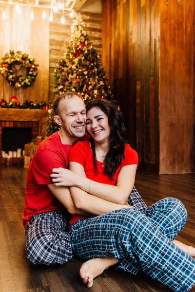 Pareja de adultos casados en camisetas rojas y pijamas abraza sentado en el suelo contra el árbol de Navidad . —  Fotos de Stock