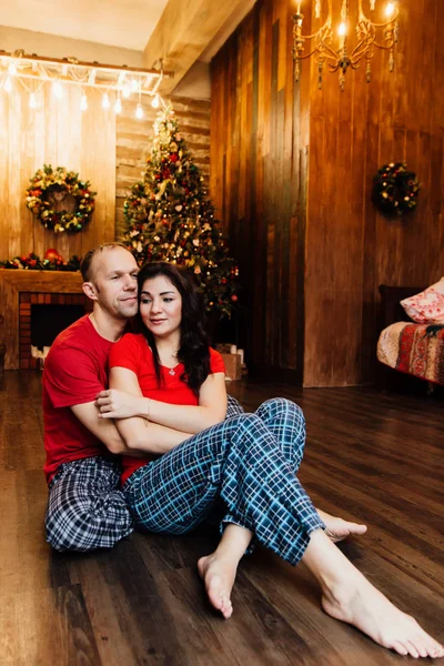 Casal adulto casado em camisetas vermelhas e pijamas abraça sentado no chão contra a árvore de Natal . — Fotografia de Stock