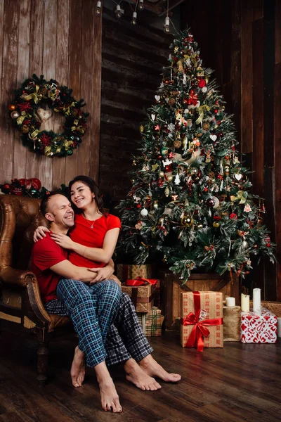 Пара в красных футболках и пижаме сидит в кресле у елки . — стоковое фото