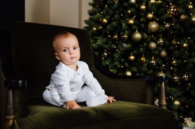 küçük çocuk Noel ağacına beyaz kaydırıcıları yeşil bir sandalyede oturur.