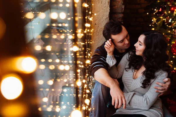 Un par de amantes abrazos sentados en el alféizar de la ventana en el estudio del loft de Navidad. chico está abrazando a la chica . —  Fotos de Stock