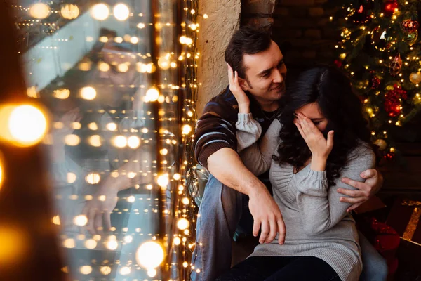 연인 커플 크리스마스 로프트 스튜디오에 창턱에 앉아 안 아. 남자는 여자를 포옹. — 스톡 사진