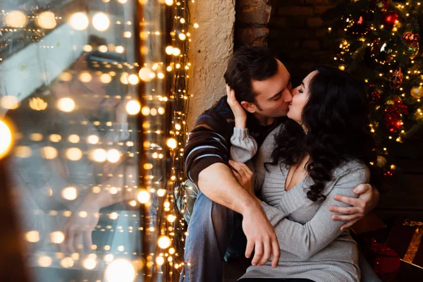 Un par de amantes abrazos sentados en el alféizar de la ventana en el estudio del loft de Navidad. chico está abrazando a la chica . —  Fotos de Stock