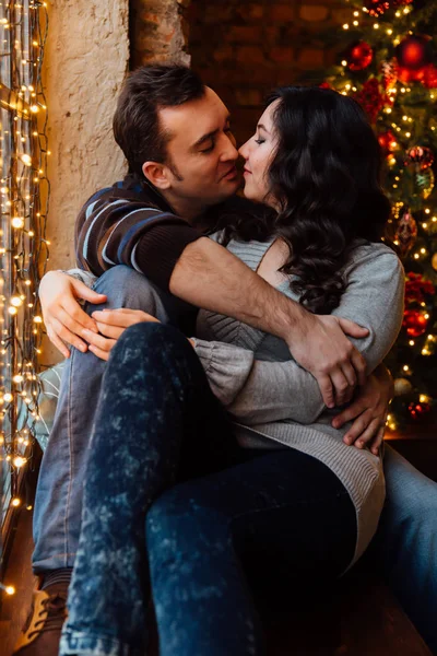 Paar liefhebbers hugs zittend op de vensterbank in de kerst loft studio. Guy is knuffelen het meisje. — Stockfoto