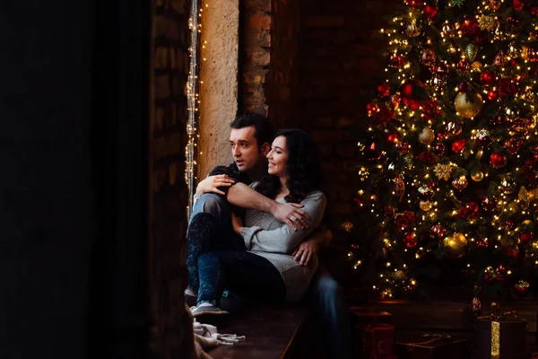 Un par de amantes abrazos sentados en el alféizar de la ventana en el estudio del loft de Navidad. chico está abrazando a la chica . —  Fotos de Stock