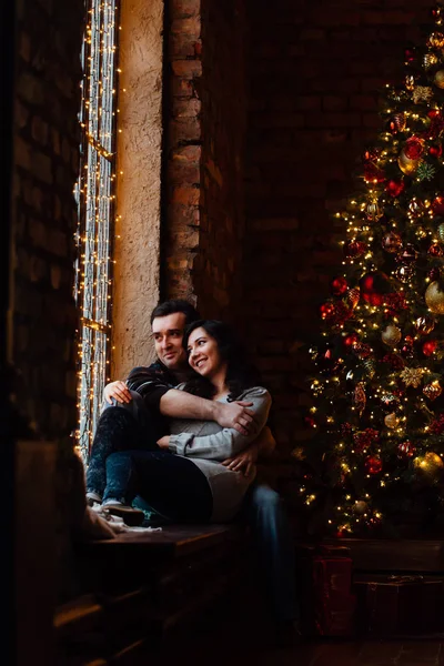 Ein Liebespaar umarmt sich auf der Fensterbank im weihnachtlichen Loft-Studio. Kerl umarmt das Mädchen. — Stockfoto