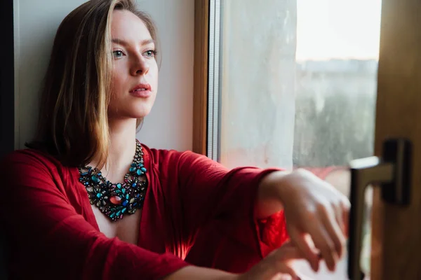 Beautiful sexy girl in a red robe is sitting on the windowsill at home. — Stock Photo, Image