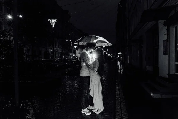 Jeune couple sous un parapluie embrasse la nuit dans une rue de la ville . — Photo