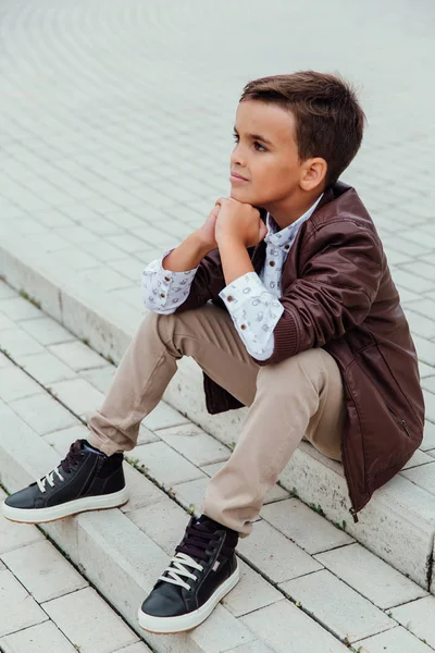 Adorable petit garçon assis dans les escaliers d'une ville, vêtu d'une veste en cuir marron . — Photo