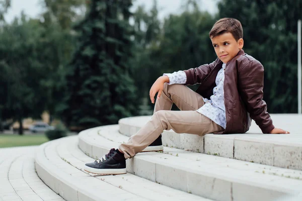 Adorable petit garçon assis dans les escaliers d'une ville, vêtu d'une veste en cuir marron . — Photo