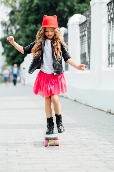 Mode élégante petite fille enfant équitation skateboard sur la rue de la ville. Chapeau rouge, veste motard noire . — Photo