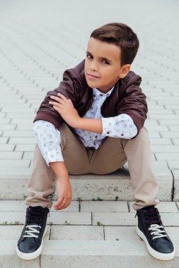 Kahverengi deri ceket giyen bir şehirde, merdivenlerde oturan sevimli küçük çocuk.