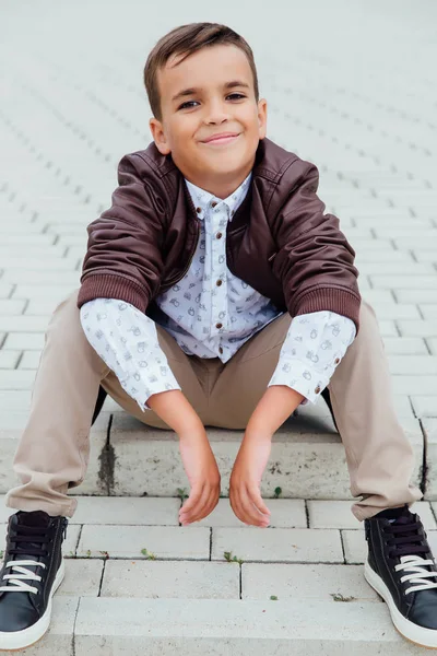 Adorable petit garçon assis dans les escaliers d'une ville, vêtu d'une veste en cuir marron . — Photo