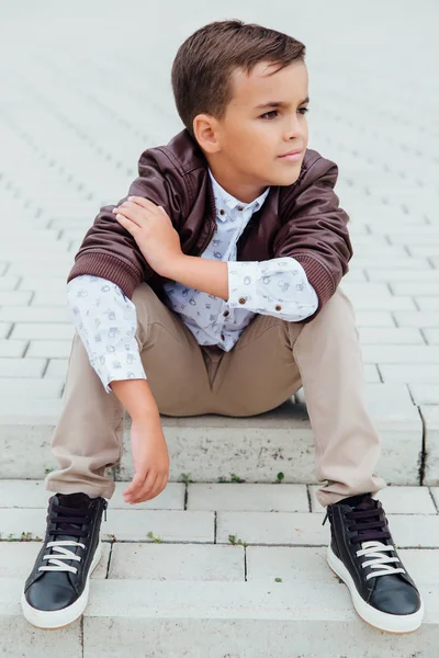 Adorable petit garçon assis dans les escaliers d'une ville, vêtu d'une veste en cuir marron . — Photo