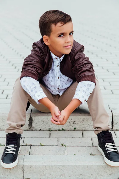 Adorable petit garçon assis dans les escaliers d'une ville, vêtu d'une veste en cuir marron . — Photo