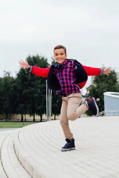 Adorabile bambino che salta in aria sulle scale di una città, indossando una giacca di pelle marrone — Foto Stock