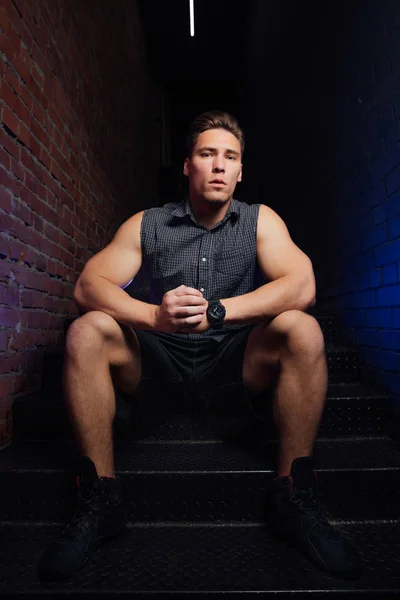 Fitness model man in shirt sleeveless posing sitting on stairs in fitness club loft. Red brick wall.