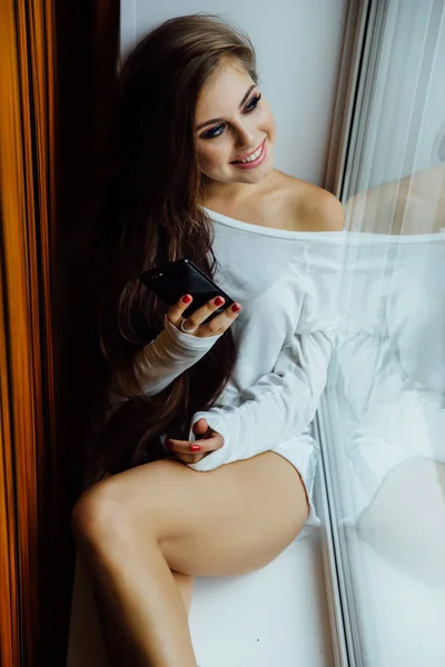 Pretty brunette girl sitting on window sill with smartphone in hands. long wavy hair. — Stock Photo, Image