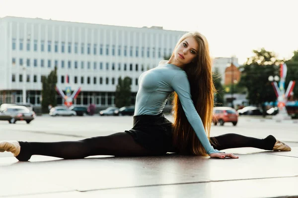 Bailarina sentada en pose gimnástica en medio de la calle de la ciudad. edificios fondo . —  Fotos de Stock