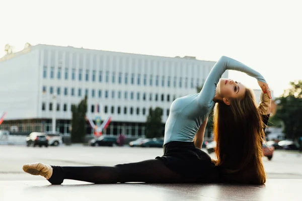 Ballerina sitting in gymnastic pose in middle of city street. buildings background. — Stock Photo, Image