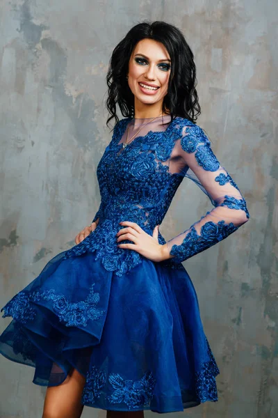 brunette woman in the blue evening dress is posing near the light texture wall background.