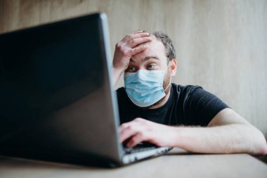 Coronavirus, sakallı yetişkin bir erkek dizüstü bilgisayarda çalışıyor. Koruyucu solunum maskesi.