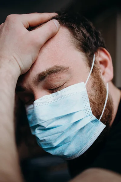 Sad balding adult man in a medical mask holding his head with his hands.