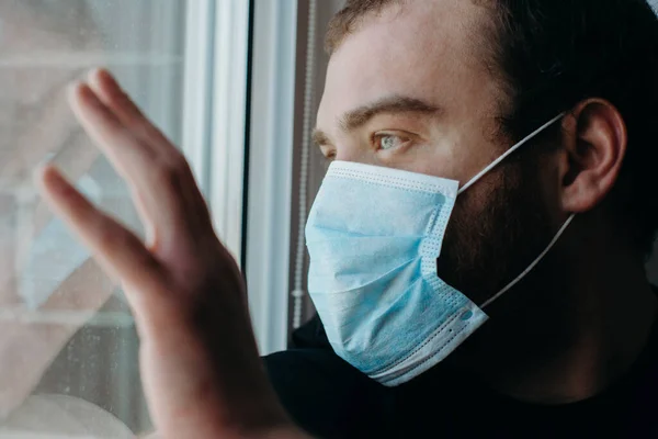 Coronavirus. Hombre enfermo de virus corona mirando a través de la ventana y usando protección antifaz . Fotos De Stock Sin Royalties Gratis