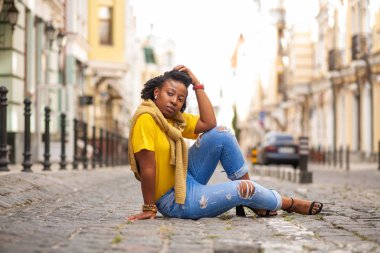 Hayat tarzı sokak kızı. Sarı tişörtlü ve kot pantolonlu Afro-Amerikalı bir kız bir şehir caddesinde duruyor. Bir fotoğraf için güzel poz veren şık bir kadın. Çok tatlı ve pozitif..