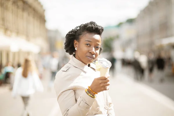 Lifestyle street girl. Beautiful and stylish black girl dressed in a tan coat on a summer street eating ice cream