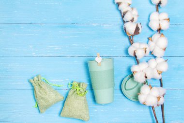 Mavi ahşap arka planda pamuk çiçekleri. Masanın üzerinde yeşil bir bardak bambu ve iki yeşil torba var. Sıfır atık.