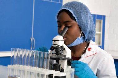 Genç Afrikalı bayan teknisyen ya da koruyucu beyaz önlüklü, şapkalı, eldivenli ve maskeli bilim adamı modern laboratuvar ya da araştırma tesisinde mikroskopla çalışıyor. Hasta biyopsisi örnekleri analiz ediliyor, kanser teşhisi konuyor.