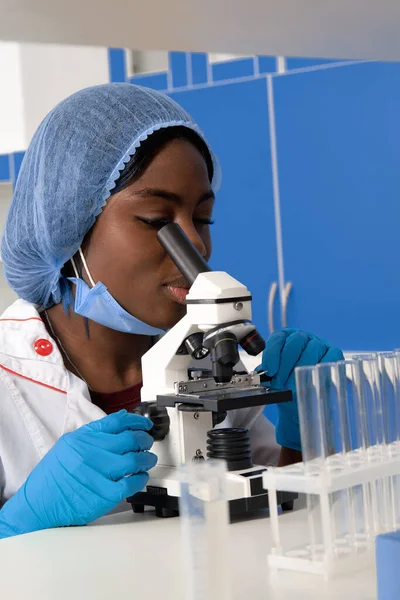 若いアフリカの女性技術者や女性科学者保護白いコートでは 手袋やマスクは 現代の研究室やテスト研究室で顕微鏡で動作します 患者の生検サンプルの分析 がんの診断 — ストック写真
